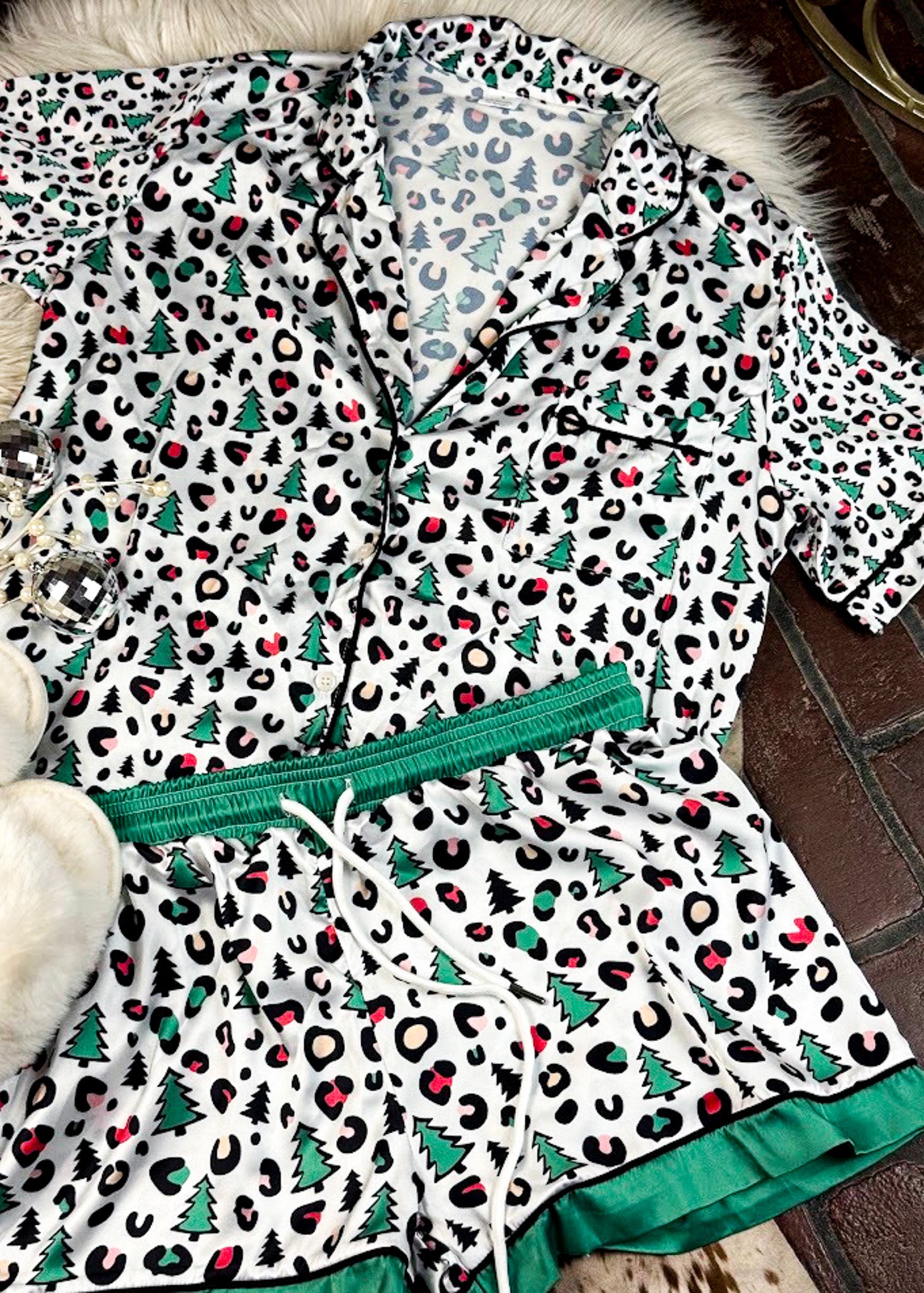 leopard print and green with christmas trees silky pajamas, two piece set with shorts, laying on a white fluffy rug with white fluffy slippers 