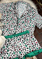 leopard print and green with christmas trees silky pajamas, two piece set with shorts, laying on a white fluffy rug with white fluffy slippers 