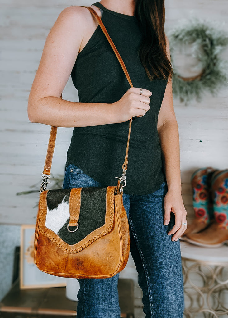 Marisol Cowhide Crossbody Handbag on model with cowboy boots and western decor in the background. handbag is leather with cowhide.