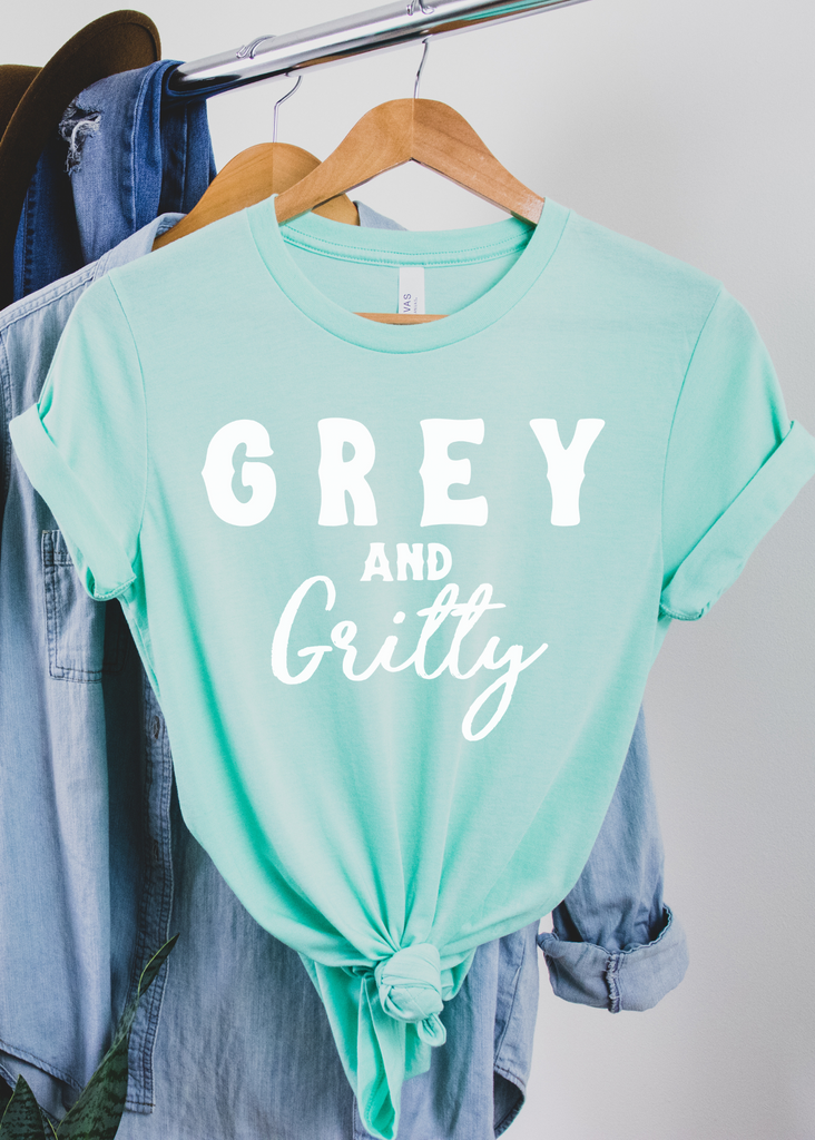 Mint short sleeve with white lettering reading "Grey and Gritty". Tee hangs from wooden hanger on a metal rack with the sleeves rolled with the bottom tied. 