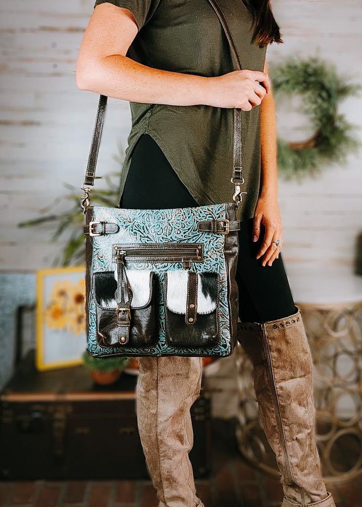 Patina Floral Cowhide Crossbody Handbag with model wearing an olive green shirt, black leggings and tan knee high boots, western decor in the background