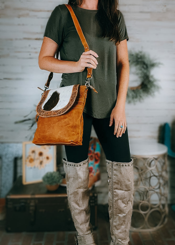 Saddleback Cowhide Crossbody Handbag on model wearing olive green shirt, black leggings and western tan knee high boots, background has western and farmhouse decor