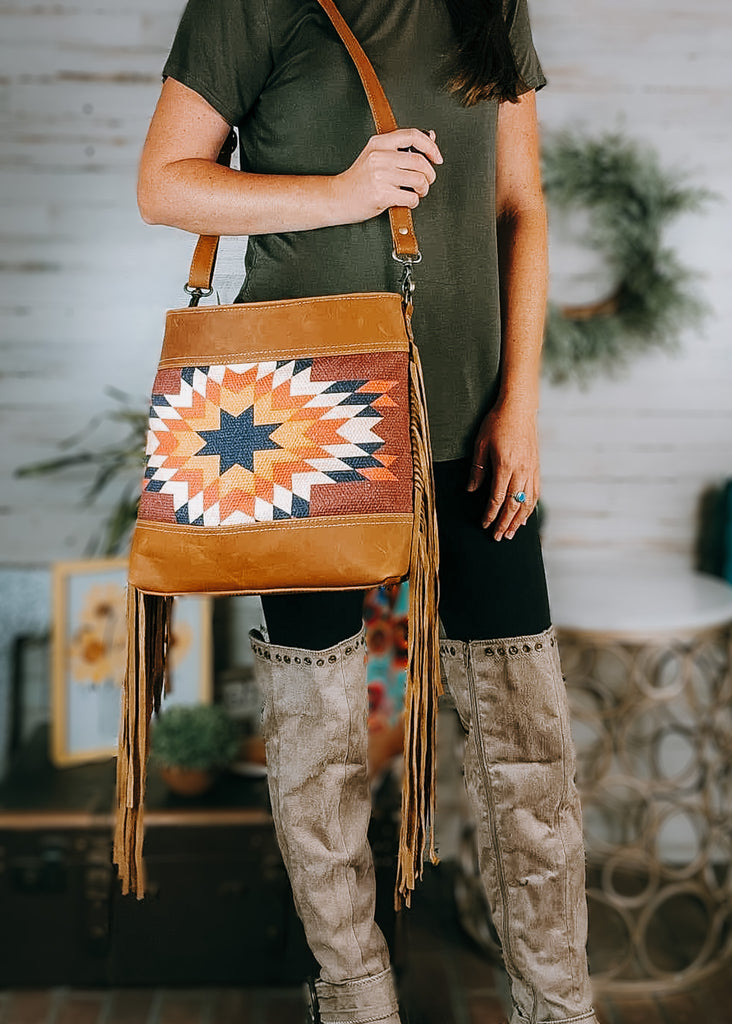 sedona star aztec crossbody handbag with model wearing an olive green shirt, black leggings and tan knee high boots, western decor in the background