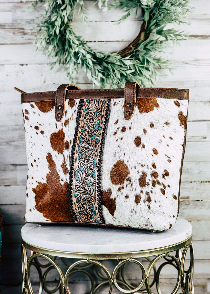 western cowhide tote handbag with teal and floral tooled leather down the center, pictured on white table with green decor
