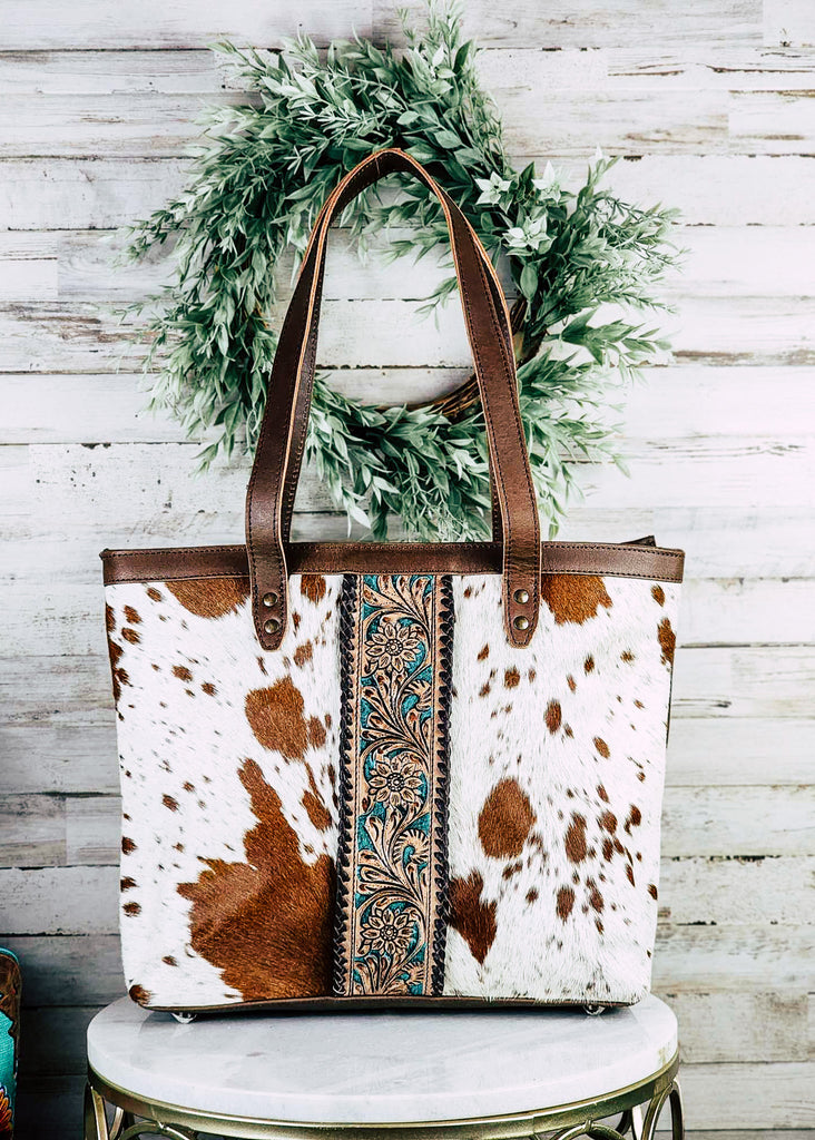 western cowhide tote handbag with teal and floral tooled leather down the center, pictured on white table with green decor