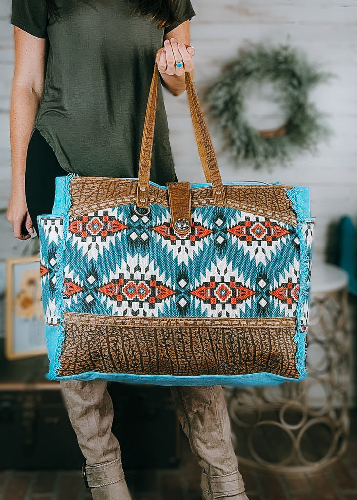 Turquoise Southwestern Weekender Bag with model wearing olive green shirt, black leggings and tan knee high boots, western decor in the background