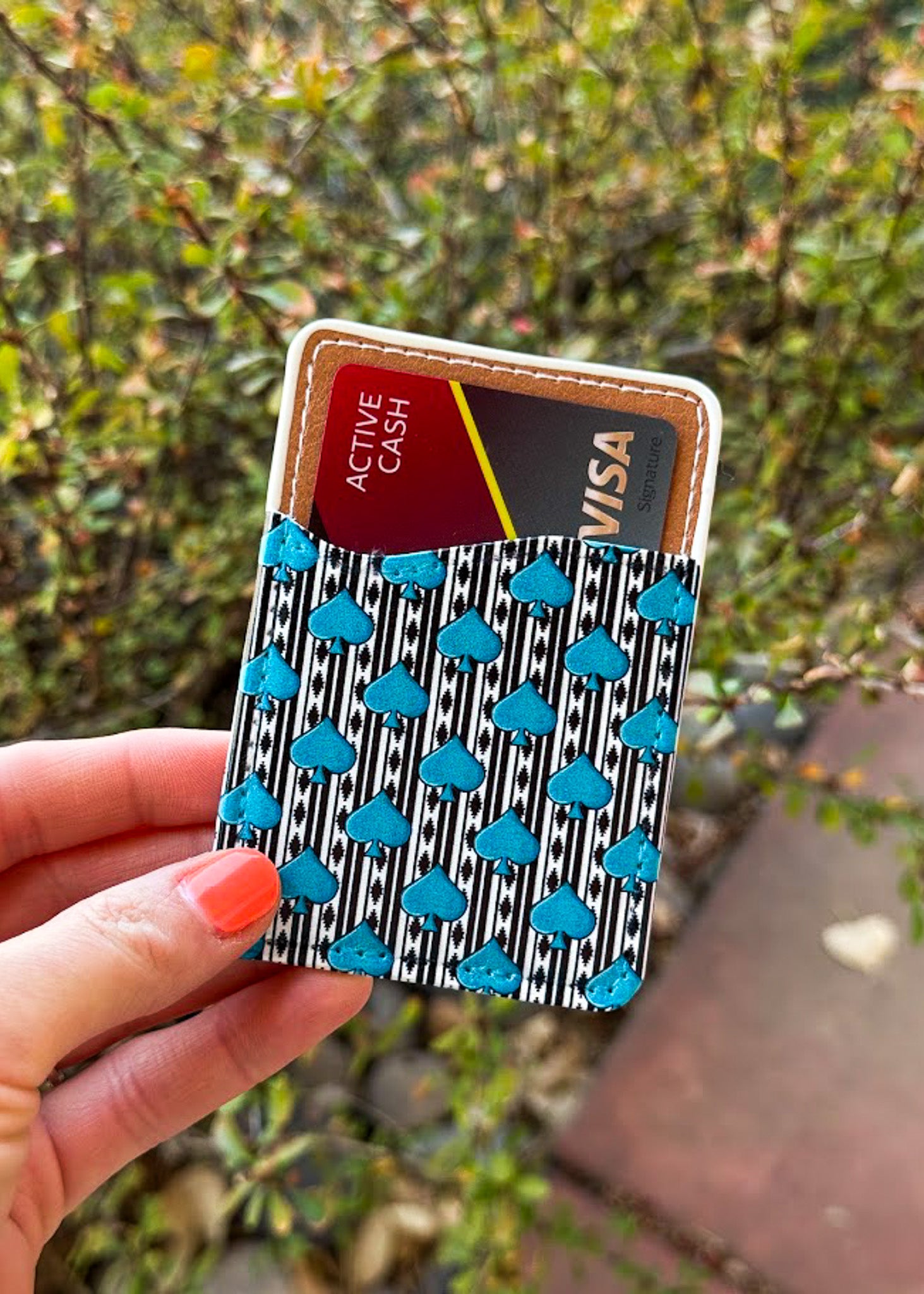 A hand holding a slim card holder wallet with a black and white striped pattern, turquoise blue cloud accents, and a tan leather trim, shown against a blurred outdoor background of green foliage.