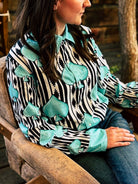 A woman wearing a long-sleeve button-up shirt with a black and white zebra stripe pattern and large turquoise spade-shaped appliques, seated in a rustic wooden chair.