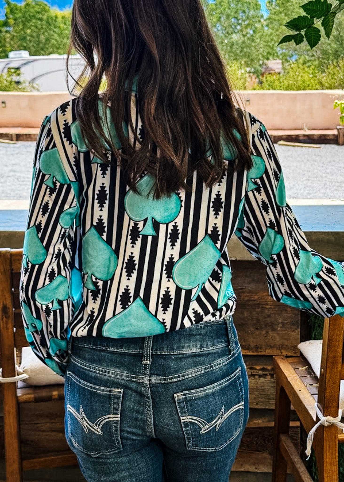 A woman viewed from behind wearing a long-sleeve blouse with a black and white vertical stripe pattern and large teal teardrop motifs, paired with blue jeans in an outdoor patio setting.