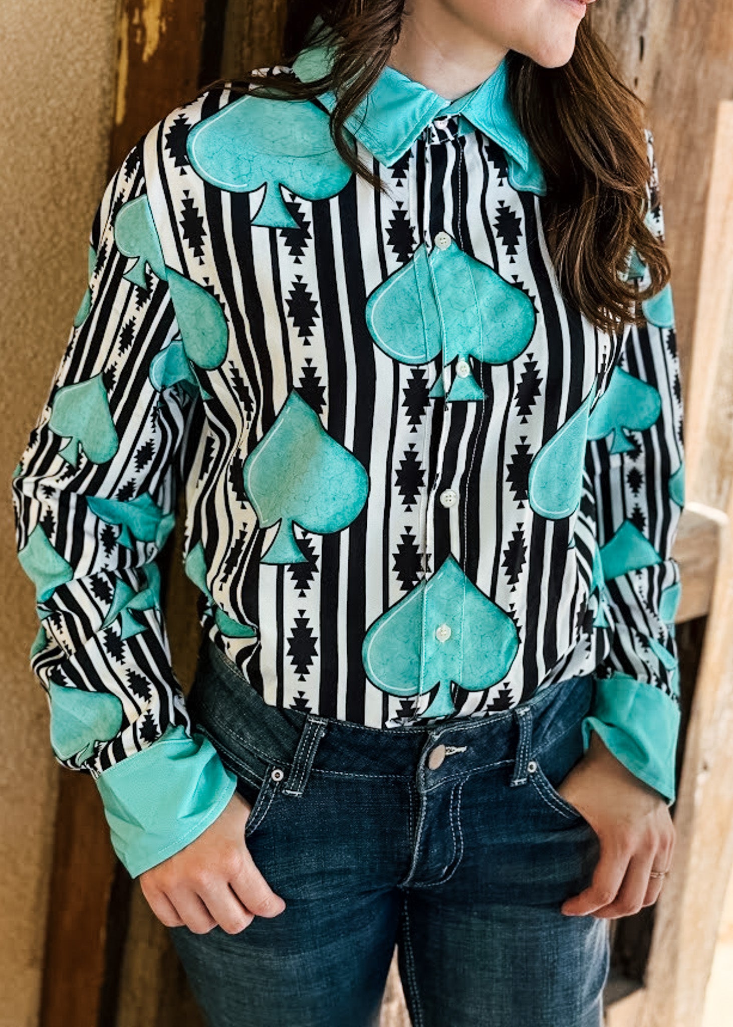 A woman wearing a long-sleeve button-up shirt with a black and white vertical stripe pattern, large teal spade motifs, and matching teal collar and cuff trims, paired with dark blue jeans.