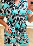 A person wearing a matching short-sleeve pajama set with a black and white vertical stripe pattern and large turquoise spade card motifs, taking a mirror selfie in a bathroom.