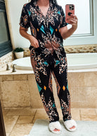 A woman takes a mirror selfie in a bathroom wearing a matching short-sleeve button-up pajama top and pants set. The set features a black base with a multicolor print combining leopard spots, geometric diamond shapes, and teal accents.