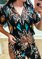 A woman wearing a matching short-sleeve button-front pajama top and shorts set with a bold abstract print. The pattern combines black, beige, brown, teal, and orange geometric shapes and leopard-like spots, photographed in a mirror selfie.