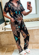 A woman takes a mirror selfie in a bathroom wearing a matching short-sleeve button-front pajama top and pants set with a black base and an abstract geometric print in beige, brown, teal, and orange. She is wearing white fluffy slide sandals.