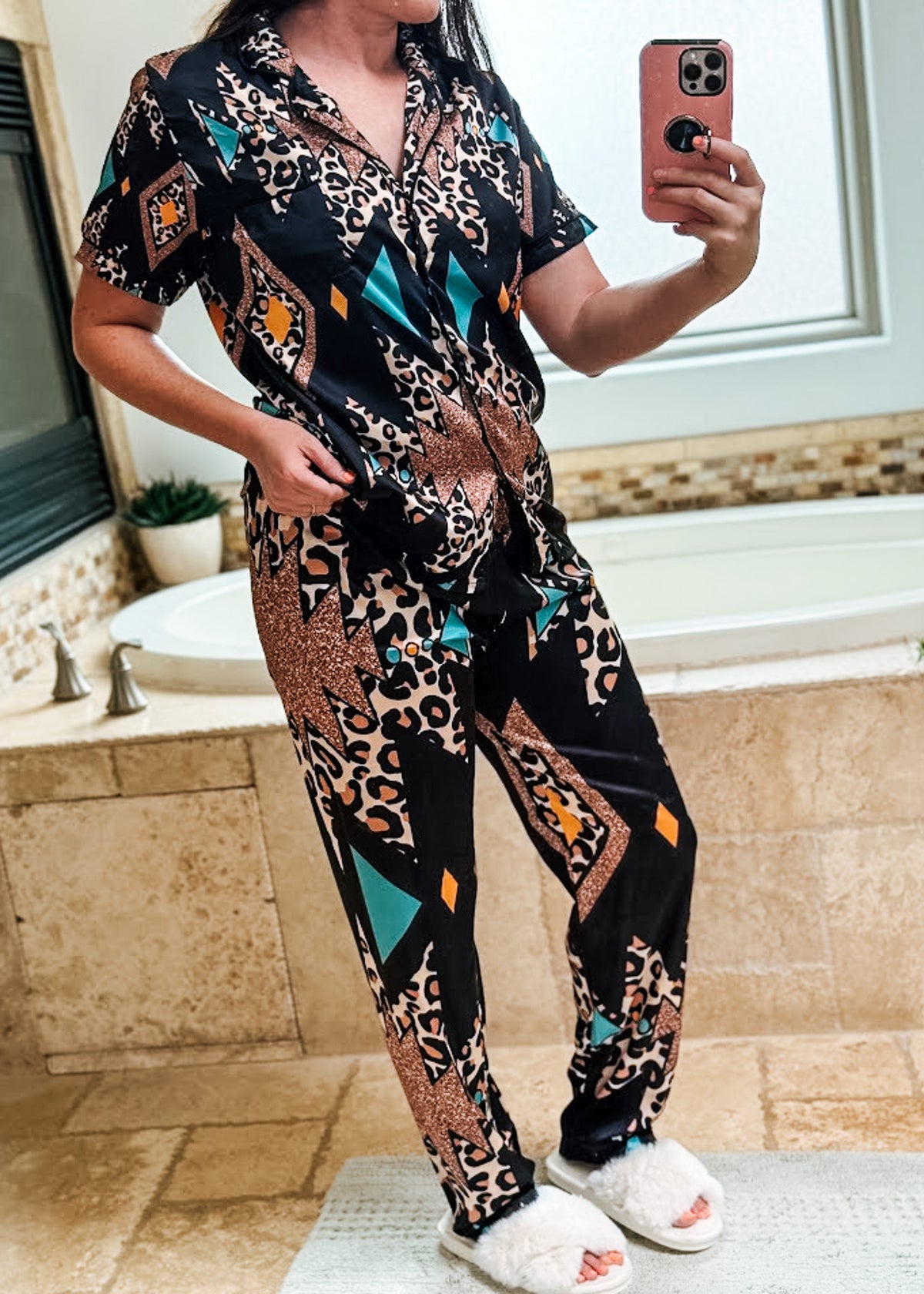 A woman takes a mirror selfie in a bathroom wearing a matching short-sleeve button-front pajama top and pants set with a black base and an abstract geometric print in beige, brown, teal, and orange. She is wearing white fluffy slide sandals.