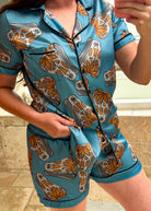 A woman wearing a matching short-sleeve button-front pajama top and shorts set in a teal blue satin-like fabric with a repeating orange, gray, and white abstract leaf or gear pattern, photographed in a mirror selfie.
