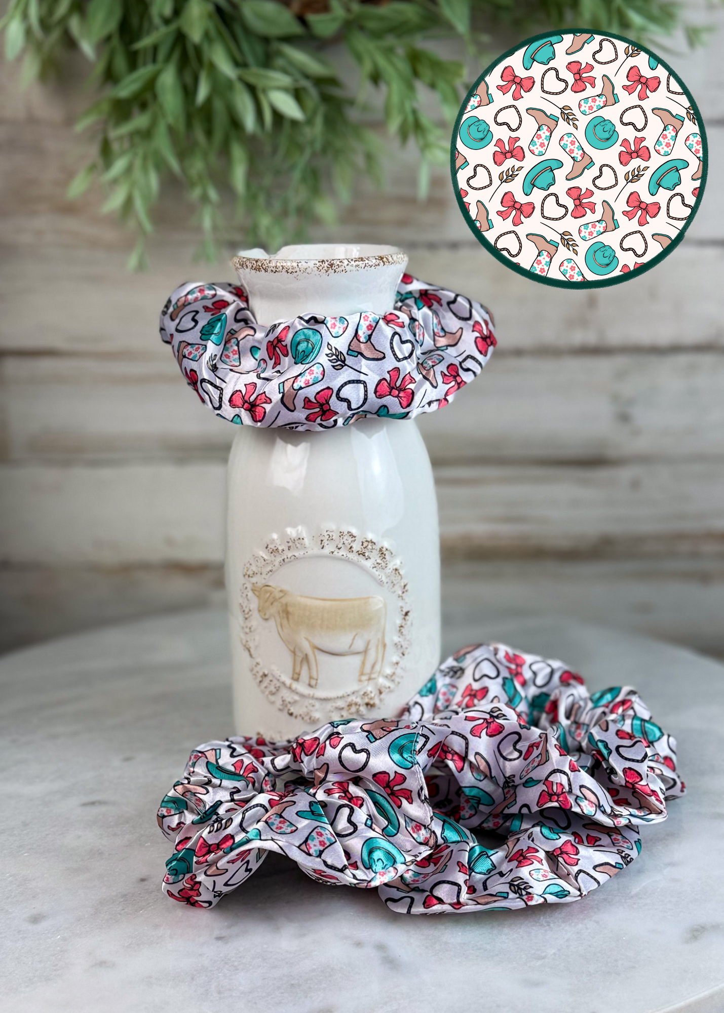 A set of fabric hair scrunchies displayed on a white ceramic bottle and on a marble surface, with a circular inset showing the detailed pattern. The scrunchies feature a white base with a colorful print of red cherries, green leaves, and black hearts.