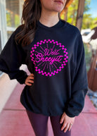 A woman wearing a black crewneck sweatshirt with a bright pink graphic that reads 'Well Sheyit' inside a pink checkerboard circle, paired with dark purple leggings, standing outdoors.