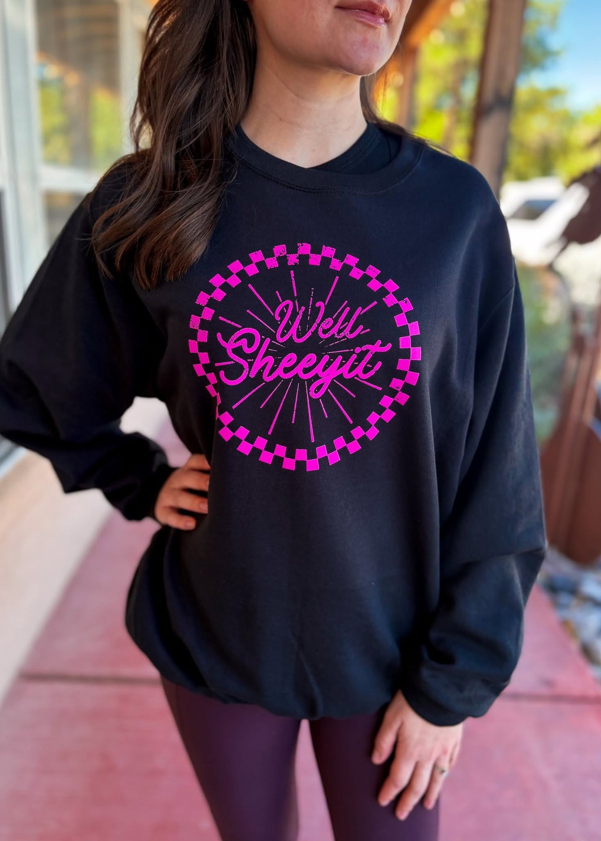 A woman wearing a black crewneck sweatshirt with a bright pink graphic that reads 'Well Sheyit' inside a pink checkerboard circle, paired with dark purple leggings, standing outdoors.