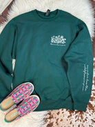 A dark green crewneck sweatshirt with a white floral graphic and text on the left chest, styled flat-lay on a white faux fur rug with a pair of pink, blue, and green patterned slip-on shoes.