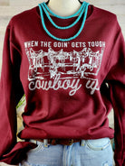A maroon long-sleeve crewneck sweatshirt with a white graphic print reading 'WHEN THE COIN' GETS TOUGH cowboy up' and an illustration of a saloon, styled on a mannequin with light blue jeans and a turquoise beaded necklace.