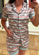 A woman takes a mirror selfie wearing a short-sleeve button-front pajama top and matching shorts with horizontal stripes in dusty pink, light blue, taupe, and white, accented with black piping and light blue trim.