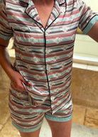 A person wearing a short-sleeve button-front pajama top and matching shorts with horizontal stripes in dusty pink, teal, brown, and white, plus a small geometric motif. The photo is cropped from the neck to the upper thighs and taken in a real indoor setting with a tiled floor and wall visible.