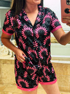 A woman wearing a short-sleeve pajama romper with a black and pink floral print, taking a mirror selfie in a bathroom setting.