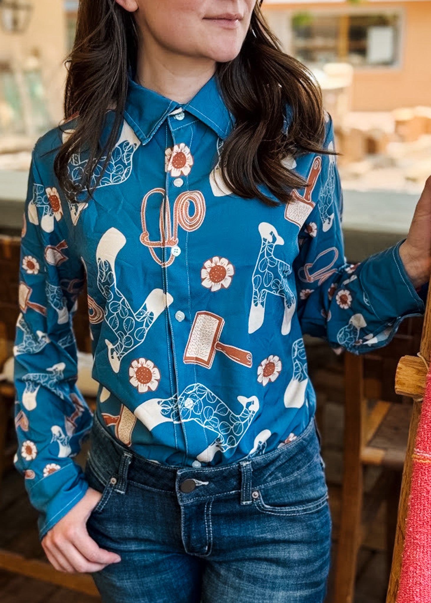 A woman wearing a long-sleeve button-up shirt with a blue base and a western-themed print featuring cowboy boots, horseshoes, flowers, and a horseshoe claw hammer. She is wearing blue jeans and standing in a rustic indoor setting.