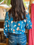 A woman viewed from behind wearing a vibrant blue long-sleeve blouse with a whimsical print of cowboy boots, mallets, and flowers, paired with blue denim jeans.