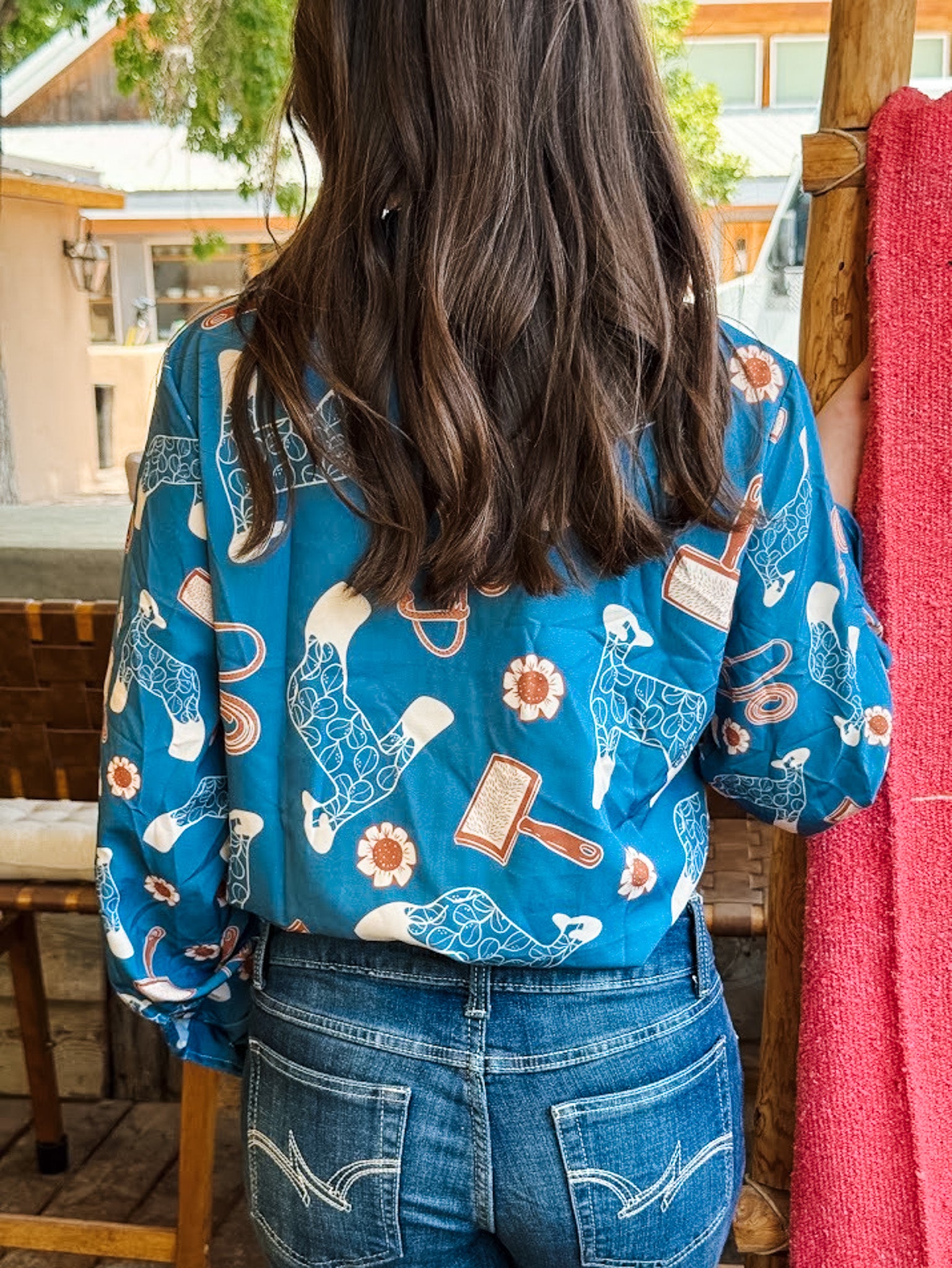 A woman viewed from behind wearing a vibrant blue long-sleeve blouse with a whimsical print of cowboy boots, mallets, and flowers, paired with blue denim jeans.