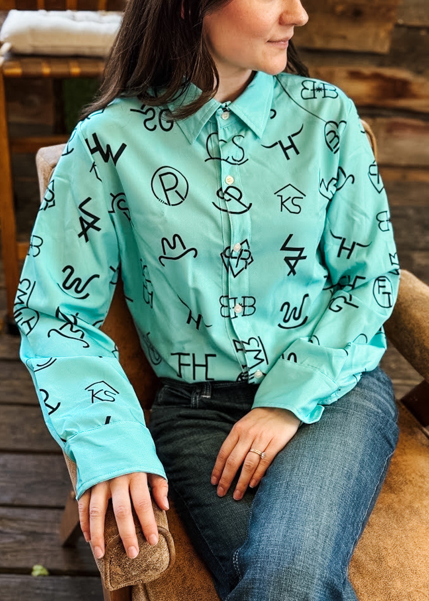 A woman wearing a mint green button-up shirt with a black abstract graphic print, paired with blue jeans, sitting on a rustic wooden chair.