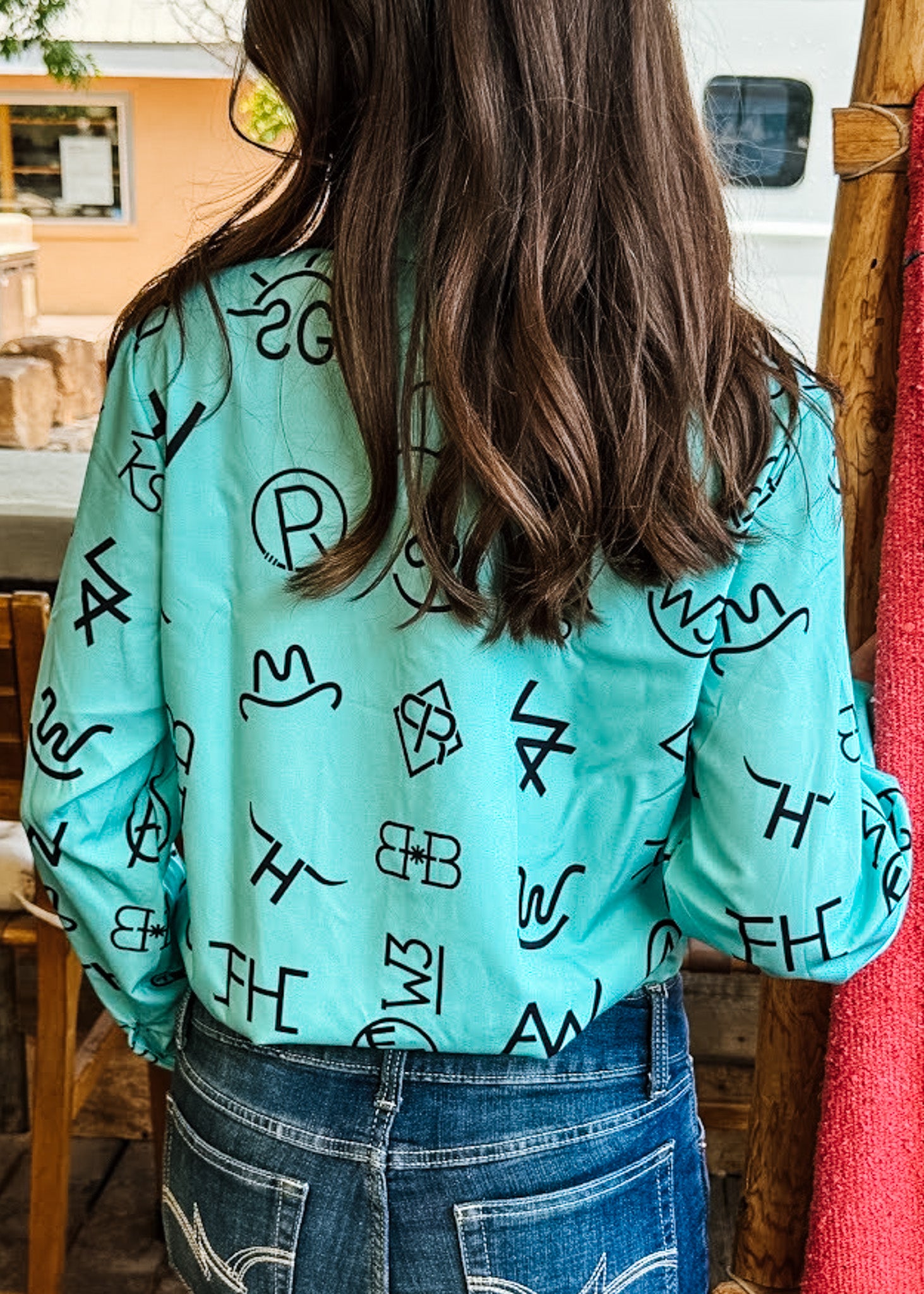 A person viewed from behind wearing a mint green long-sleeve button-up shirt with a black abstract graphic print, paired with blue jeans in an outdoor rustic setting.