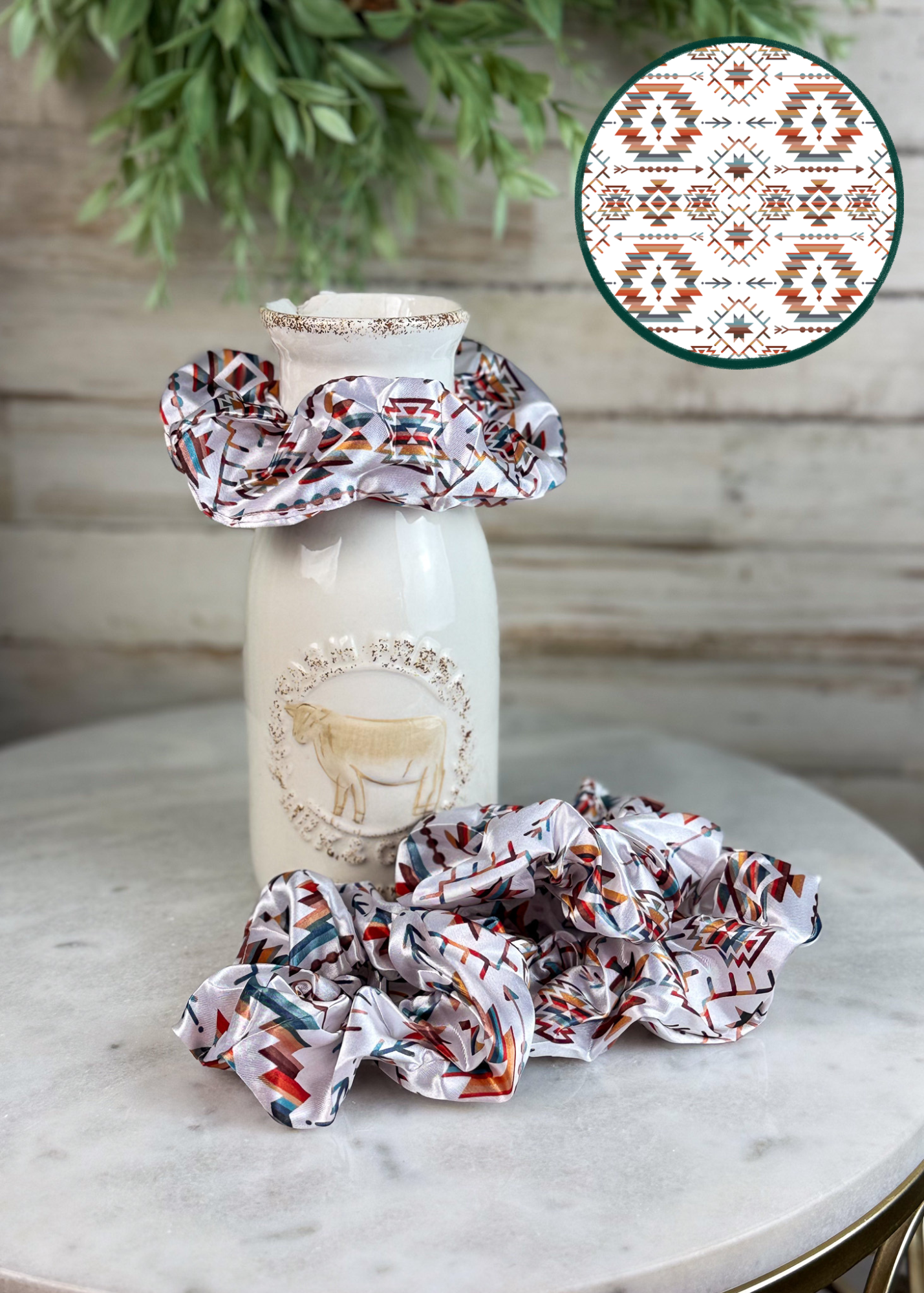 Three fabric scrunchies with a Southwestern geometric pattern in red, orange, and teal on a white background are displayed on a ceramic milk bottle and a marble surface. A circular inset in the top right shows a close-up of the pattern.