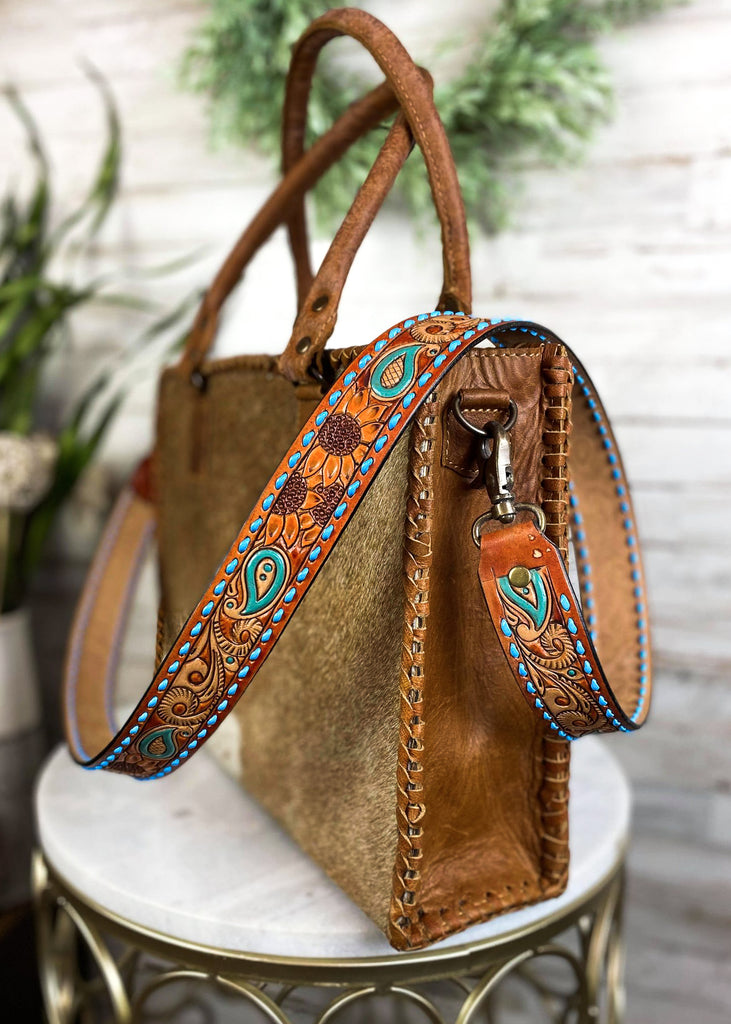 Cowgirl purse strap with sunflower tooling design. Blue buck stitch on the side of the strap, sunflowers and paisley tooling with teal accents. Strap is attached to cowhide purse sitting on white marble stool.