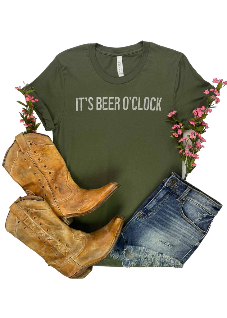 CLOSEOUT-Army green short sleeve tee with off white lettering reading "It's Beer O'Clock". Tee is laid on white background with pink floral decor, jean shorts, and boots. 