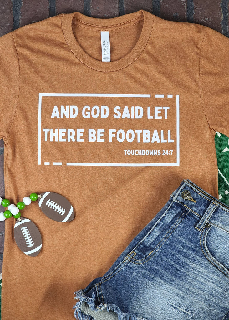 Autumn (burnt orange) short sleeve tee with white box outline and lettering that reads "And god said let there be football -touchdowns 24:7" laying on a brick background. Jean shorts lay on top of shirt with football decorations around shirt. 