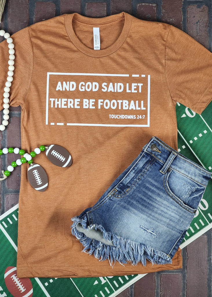 Autumn (burnt orange) short sleeve tee with white box outline and lettering that reads "And god said let there be football -touchdowns 24:7" laying on a brick background. Jean shorts lay on top of shirt with football decorations around shirt. 