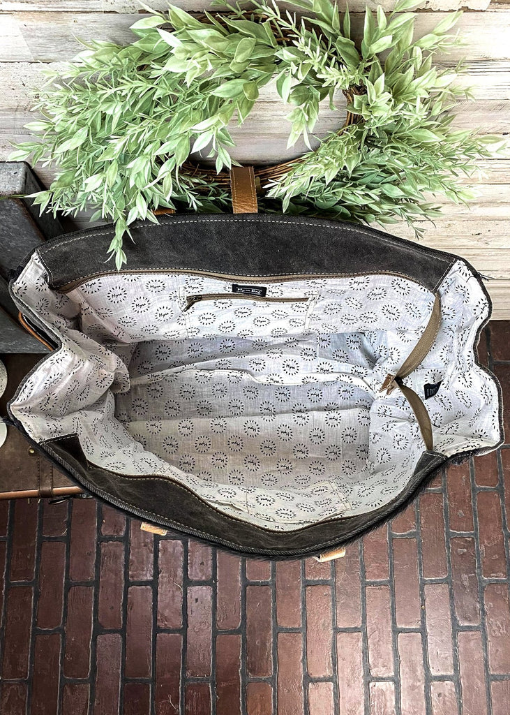 Inside view of Black and white cowhide weekender handbag with brown leather floral tooled accents at the bottom with brown leather shoulder straps, with top zip closure and flap over snap button closure, with canvas backing and back zipper pocket, with inside zipper pocket and two open inside pockets, taken inside on white table with green floral décor