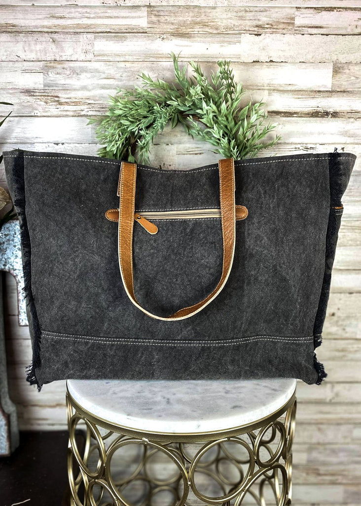 Back view of Black and white cowhide weekender handbag with brown leather floral tooled accents at the bottom with brown leather shoulder straps, with top zip closure and flap over snap button closure, with canvas backing and back zipper pocket, with inside zipper pocket and two open inside pockets, taken inside on white table with green floral décor
