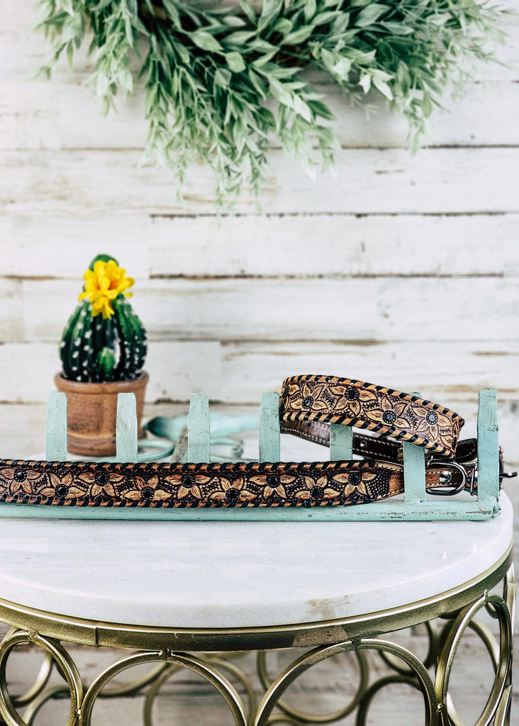 Brown floral tooled dog collar with darker contrasting whip stitch. Collars lay on marble table one is wrapped around a blue metal decor rake and one lays stretched along the side of the rake. 