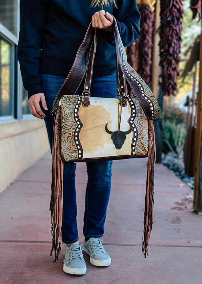 Western crossbody bag with cowhide inlay with tooled leather side panels. White whip stitch lines the leather panels. Steer skull cutout is attached to handle straps. Fringe hangs from the side of the bag. Model wears this over her shoulder with matching tooled and whip stitch shoulder strap. Style B