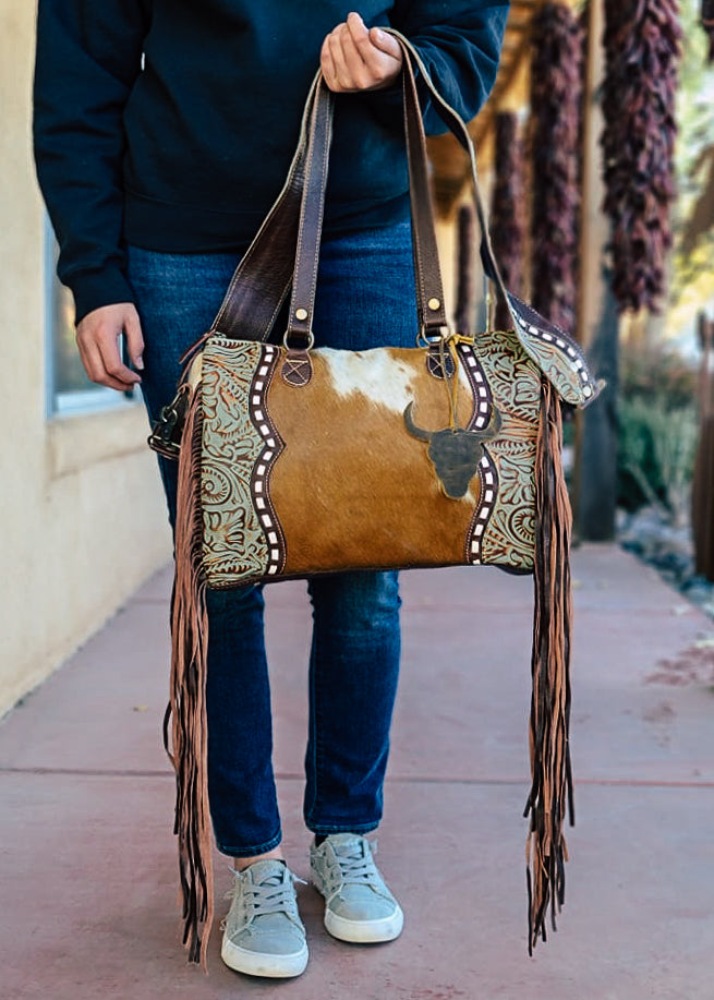 Western crossbody bag with cowhide inlay with tooled leather side panels. White whip stitch lines the leather panels. Steer skull cutout is attached to handle straps. Fringe hangs from the side of the bag. Model wears this over her shoulder with matching tooled and whip stitch shoulder strap.Style B
