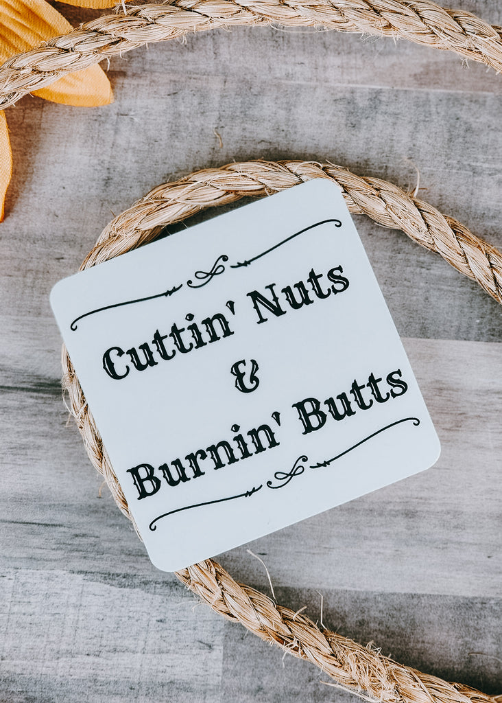 Rounded square sticker with black design and lettering reading "Cuttin' Nuts and Burnin" Butts". Sticker lays on rope on wooden table. 