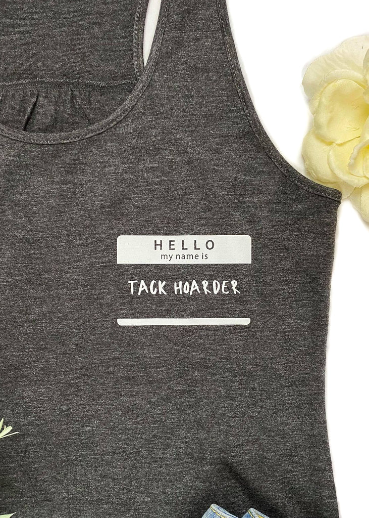 Charcoal colored tank top with name tag graphic in the right (left on person) that says "Hello my name is Tack Hoarder" in white ink, laid flat on white surface with denim shorts, cowgirl hat, and turquoise necklace, with green floral décor 