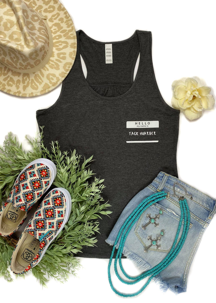 Charcoal colored tank top with name tag graphic in the right (left on person) that says "Hello my name is Tack Hoarder" in white ink, laid flat on white surface with denim shorts, cowgirl hat, and turquoise necklace, with green floral décor 