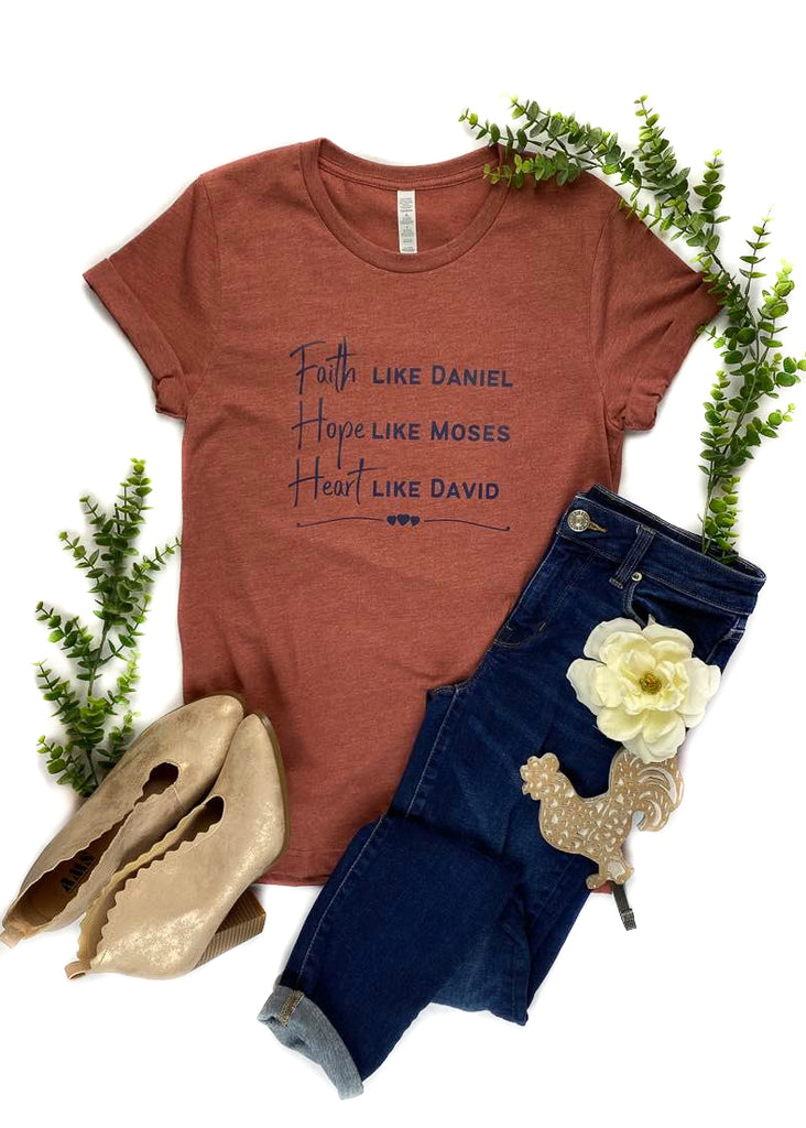Clay colored short sleeve graphic tee with "Faith Like David, Hope Like Moses, Heart Like David" and three small hearts in the middle of tee, laid flat on white surface with floral décor 