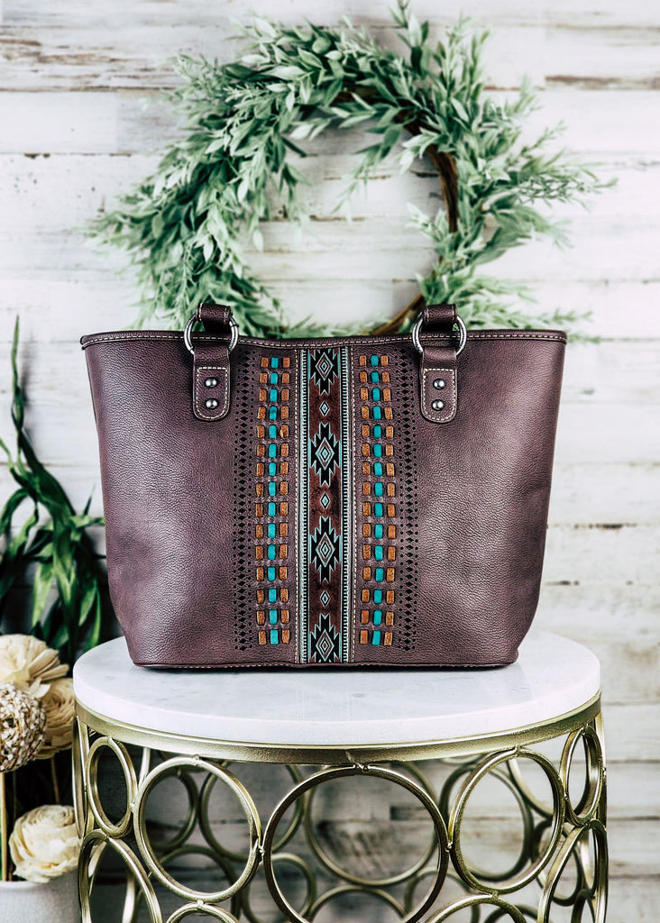 Coffee concealed carry handbag with teal and caramel brown whip stitch running down the centered Aztec teal embossed design. Purse is displayed on marble table. 