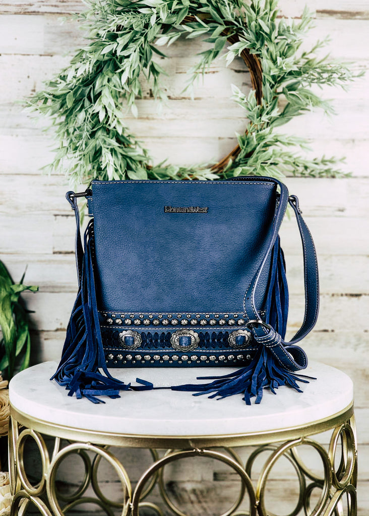 Concealed carry navy leather with accent silver conchos and studs. Blue accented stitching with silver concho buckles. Navy fringe hangs from the side of the purse with adjustable shoulder strap. Purse sits on marble table.