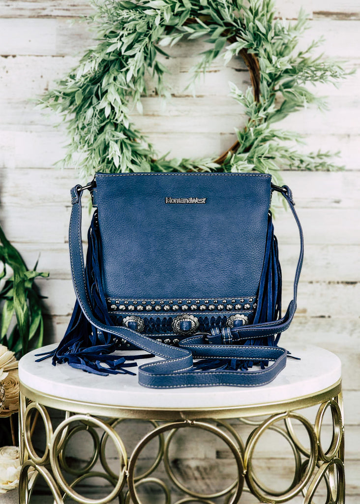 Concealed carry navy leather with accent silver conchos and studs. Blue accented stitching with silver concho buckles. Navy fringe hangs from the side of the purse with adjustable shoulder strap. Purse is displayed on a marble table top. 